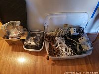 Wide view of plastic bin with power strips, extension cords, and other hardware items on wooden floor.