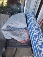Photo showing both the gray quilted moving blanket and the blue and white plastic outdoor rug placed on an outdoor table and partially rolled out to show pattern and texture.