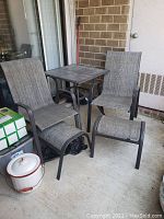Patio set on balcony showing two high-back chairs with matching ottomans and tile top table arranged on concrete floor.