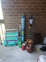 Beach chair, umbrella, watering can, planters, decorative lamp, and wicker shelf stand arranged against brick wall on balcony.