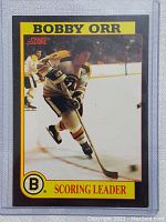 Front of the card showing Bobby Orr skating with puck in Boston Bruins jersey on ice rink with Score and Scoring Leader text.