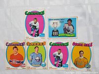 Photo showing six NHL vintage 1971 hockey cards featuring players with team names: Dan Johnson (Canucks), Gilbert Perreault (Calder Trophy), Pete Mahovlich (Canadiens), Phil Goyette (Sabres), Dan Johnson (Canucks duplicate), Paul Shmyr (Golden Seals). Colorful backgrounds and team logos visible.