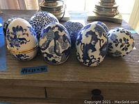 Front view of all 8 ceramic eggs and spheres displayed on wooden surface, showing varied blue patterns on white backgrounds, one with visible hinge.