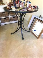 Full view of round table showing glass top with various decorative items and metal frame base with scroll details, placed on carpet.
