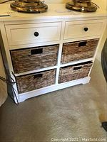 White storage unit with two drawers and four pull-out wicker baskets, shown from front angle with lamps on top, placed on carpeted floor.