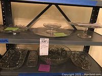 Full view of vintage glassware on metal shelving, showing cake stand and various serving dishes.