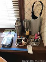 Full view of wine thermos, cocktail shaker, insulated wine goblet, insulated wine bottle caddy with rhinestones and black strap, and bar tools on wooden surface near window