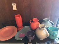 Full view of nine pieces of Fiesta Ware including pitchers, plates, mugs, and a tall orange vase or pitcher in various colors such as orange-red, light blue, cream, turquoise, and purple.