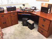 Full view of the three piece sectional wooden corner desk with electronics on top. Shows two drawer units, cupboard doors and flat work surfaces.