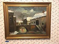 Framed oil painting showing a winter landscape scene with a stone bridge, old buildings covered in snow, and a cloudy sky.