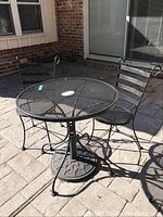 Full view of the patio set outdoors showing one round metal mesh table with two matching chairs on either side in a paved patio area.