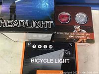Photo of two boxed bicycle light kits. One box labeled Headlight shows a silhouette cyclist against a night sky. The other box is labeled Pagao Drive Pro Bicycle Light Kits showing front and rear lights.
