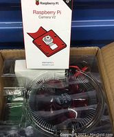 Photo showing Raspberry Pi Camera V2 box, microchip board with large circular cooling fan, and wiring, all inside cardboard box.
