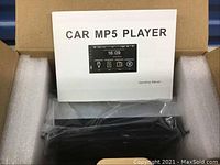 Photo of the open box containing the black car MP5 player and an operating manual labeled 'CAR MP5 PLAYER'.