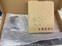 Ring light set inside an open brown cardboard box with bubble wrap covering parts, including a desk clamp and cables.