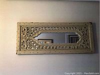 Photo shows rectangular antique mirror with distressed cream carved wooden frame and floral designs, hanging on wall.