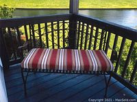 Outdoor metal bench with decorative scrollwork backrest and striped cushion, photographed on a porch overlooking a pond.
