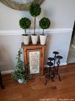 Overview of wood cabinet with floral painting and three faux topiary plants on top, faux ivy to left, and grouped four iron candle holders to right on hardwood floor