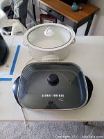 George Foreman electric skillet rectangular with glass lid and black knob, front view showing logo on cooking surface, Rival Crock Pot slow cooker round white with glass lid in background.