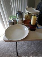 View of various pitchers, bowls, canisters, candle and holder arranged on white table.