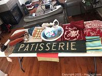 Wide view of the lot items on table showing linens, wash basin, creamer, sugar bowl, iron, and wall sign