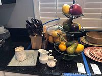 Overview of lot items on kitchen counter including cutlery block with knives, ceramic candle holder, soap dispenser, creamers, decorative plates, tiered fruit stand and bowl with faux fruit.