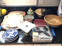 Overview photo showing multiple ceramic bowls, decorative plates, corkscrew, pedestal dish, and serving trays on counter.