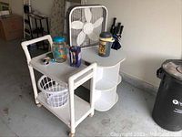 Photo showing white plastic laundry carts with baskets, Holmes 20x20 electric box fan, small white table with curved shelves, iced tea dispenser, other miscellaneous household items on concrete floor.