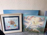 Photo showing three beach scene oils and one shadow box three-dimensional artwork leaning against wall