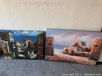 Photo showing two paintings side by side against a wall on the floor. Left painting is the Mediterranean village scene, right is the Pueblo adobe buildings scene signed Thomas.