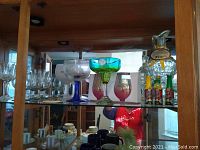 Display shelf with various specialty glasses including Cuban cafe bongo glasses, Amicl margarita glass, clear wine glasses, two decorative bottled glass containers, and boxed pink rose wine glasses