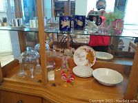 Overview of wooden display cabinet with glass doors showing the Zodiac coffee mugs and white plates and bowls on bottom shelf, and the espresso cups on upper shelf.