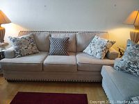 Front view of beige fabric three-seat sofa with four throw pillows, nailhead trim