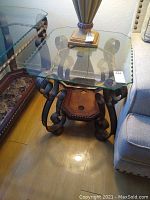 End table with beveled glass top showing scalloped edges, wood and wrought iron sculpted base with scroll details and lower wooden shelf missing the marble insert.