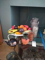 Overview of the lot including white plastic bowl with colorful faux fruit, black Asian teapot, two vases, metal milk can and glass jar on glass table.
