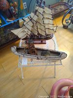 Photo of vintage sailboat model with fabric sails and rigging, canoe model with seated figure, both placed on the small white metal lattice table.