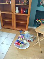 Photo showing assorted vases and other items arranged on the floor near two wooden shelves and a small foldable wooden table.
