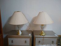 Pair of brass colored lamps with cream fabric shades placed on silver dressers.