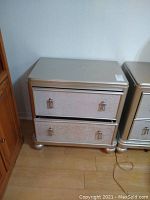 Front view of the silver finish night stand showing two drawers with silver handles and rounded feet.