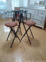 Three black metal bar stools with collapsible frames and round brown faux suede seats arranged on light wood flooring.