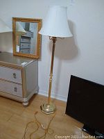 Full view of brass floor lamp standing next to a small dresser and mirror, showing the overall size and condition including lamp cord with switch.