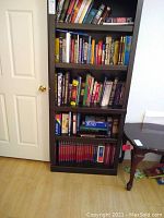 Full view of the dark composite wood bookcase filled with various books on all five shelves, against a white wall next to a door and small table with some books on the floor.