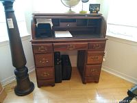 Front view of brown roll top desk showing seven drawers, roll top open with compartments inside, and two briefcase style cases placed underneath.
