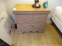 Full front view of light wood grain composite night stand with two drawers, metal handles, decorative scalloped base, wear on top surface.