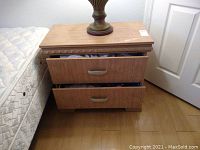 Night stand placed next to a mattress showing two drawers partially open and table lamp on top indicating size and details