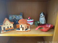 Two clay houses on a wooden shelf, one with steep roof and painted windows, the other round with terracotta tiles and flower decorations, along with blue bear lodge and other items.