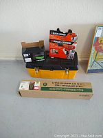 Overview of toolbox with drill box, drill bits, and green wire tie box on a floor against white wall