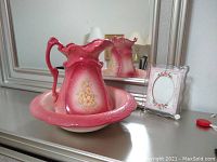 Pink reproduction ceramic wash basin and matching pitcher with floral decoration, shown on a dresser in front of a mirror, also includes a rectangular pink and white florally decorated picture frame.