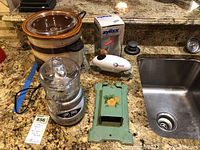 Photo showing all five items together on kitchen counter: Rival crock pot, Cuisinart mini prep chopper, Zyliss hand chopper, One Touch jar opener, and antique match storage container.