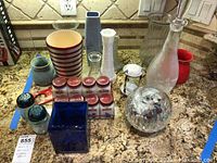 Photo showing nine milk glass spice jars with red lids and labels in a rusted metal rack, blue cobalt planter, striped ceramic pot, and other items on granite kitchen countertop.